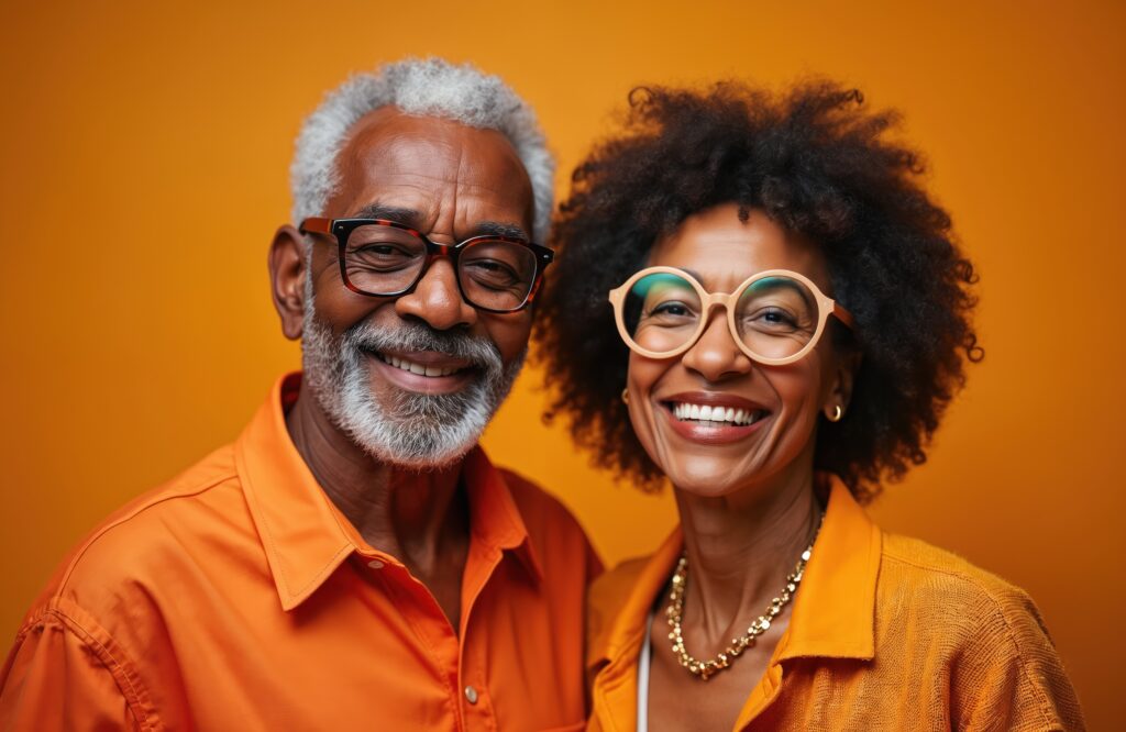 Portrait of smiling senior African American couple wearing modern orange clothes, glasses against orange background. Elderly man, woman enjoy retirement, active lifestyle. 